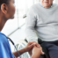 Healthcare worker in scrubs holds an elderly man’s hand while speaking with him as he sits in a wheelchair in a bright clinic setting.